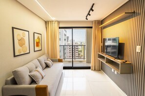 A modern living room with a sofa, a television on a stand, and a large window with a view of buildings. - Move #801 - Apartment With Balcony by Carpediem (João Pessoa)
