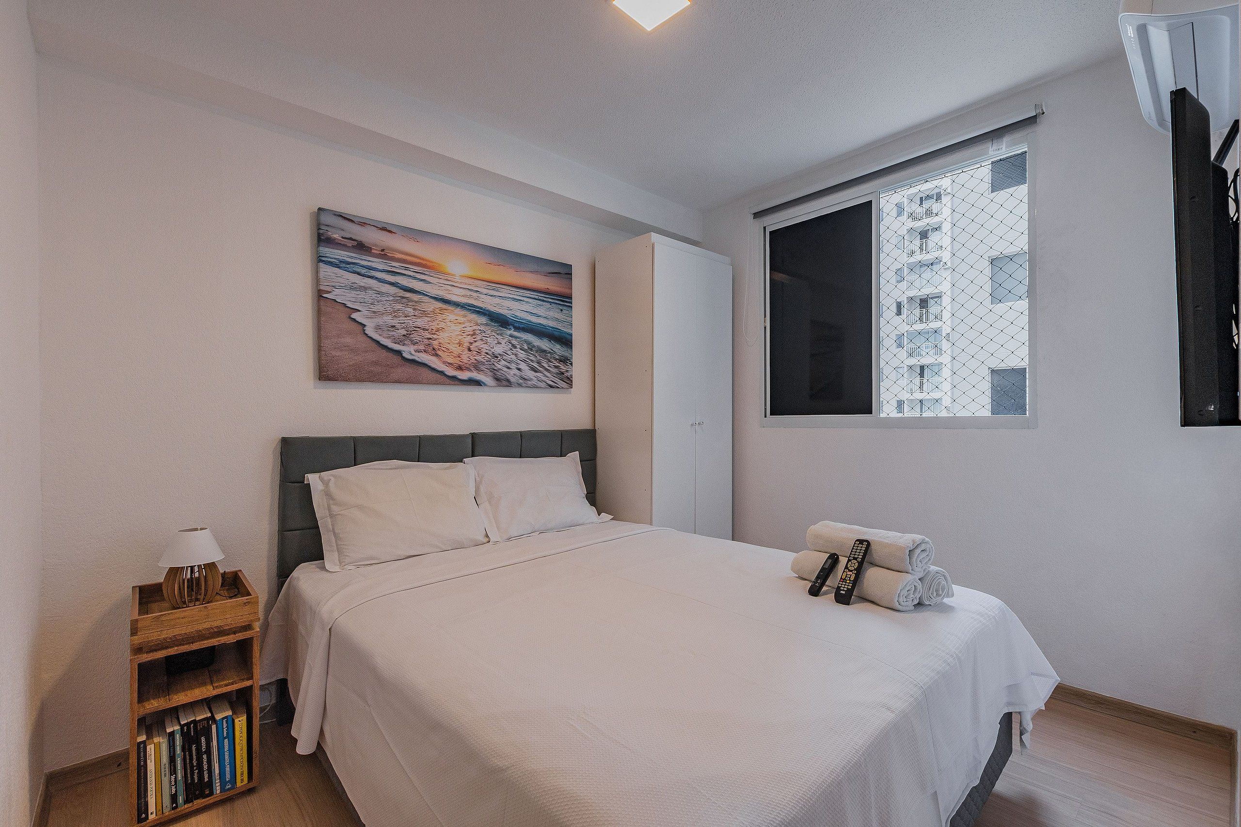 Un dormitorio con cama, mesita de noche, estantería, un cuadro, una ventana con vista a un edificio y una puerta.