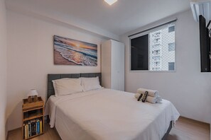 Ein Schlafzimmer mit einem Bett, einem Nachttisch, einem Bücherregal, einem Gemälde, einem Fenster mit Blick auf ein Gebäude und einer Tür.