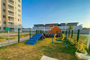 A playground with a slide and tire swings, surrounded by a fence, with a building in the background. - Mirante #1108 - Apartment in Fortaleza by Carpediem (Fortaleza)