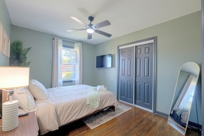 Quarto com cama, ventilador de teto, televisão, porta e janela com cortinas.