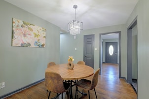 A dining area with a round table, chairs, a vase with flowers, a ceiling light fixture, and a door in the background.