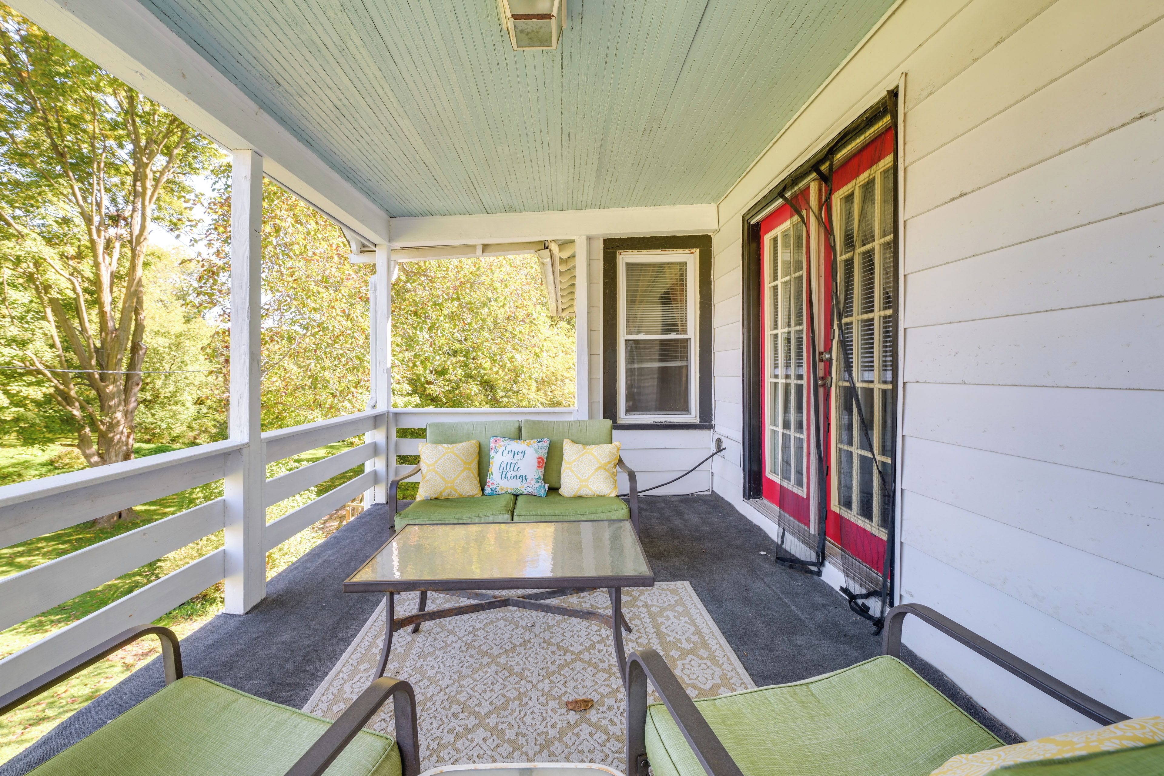 A covered porch with a green cushioned bench, a small table, and a view of trees.