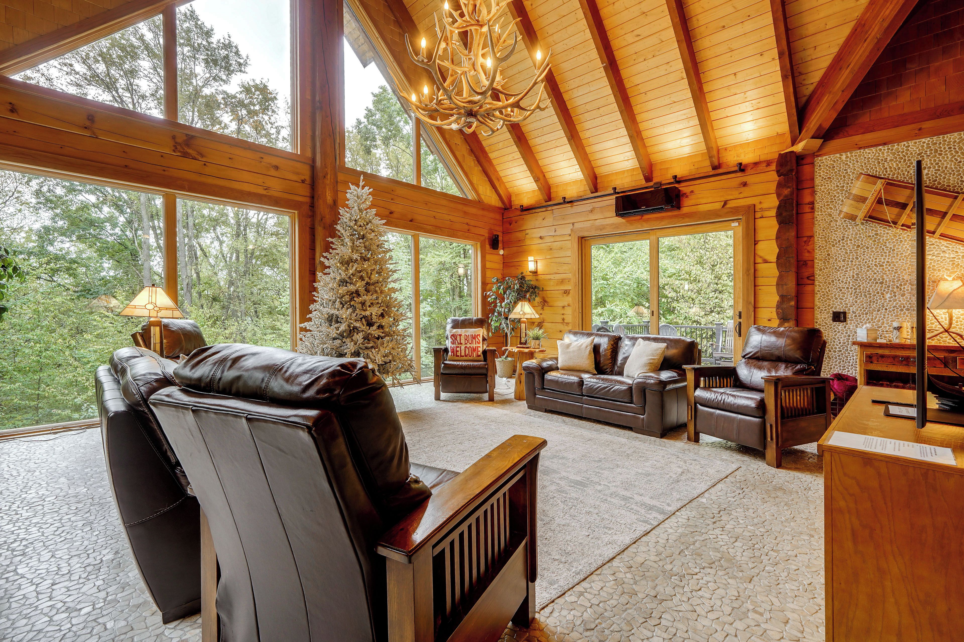 A log cabin interior with a fireplace, leather sofas, a Christmas tree, and large windows.