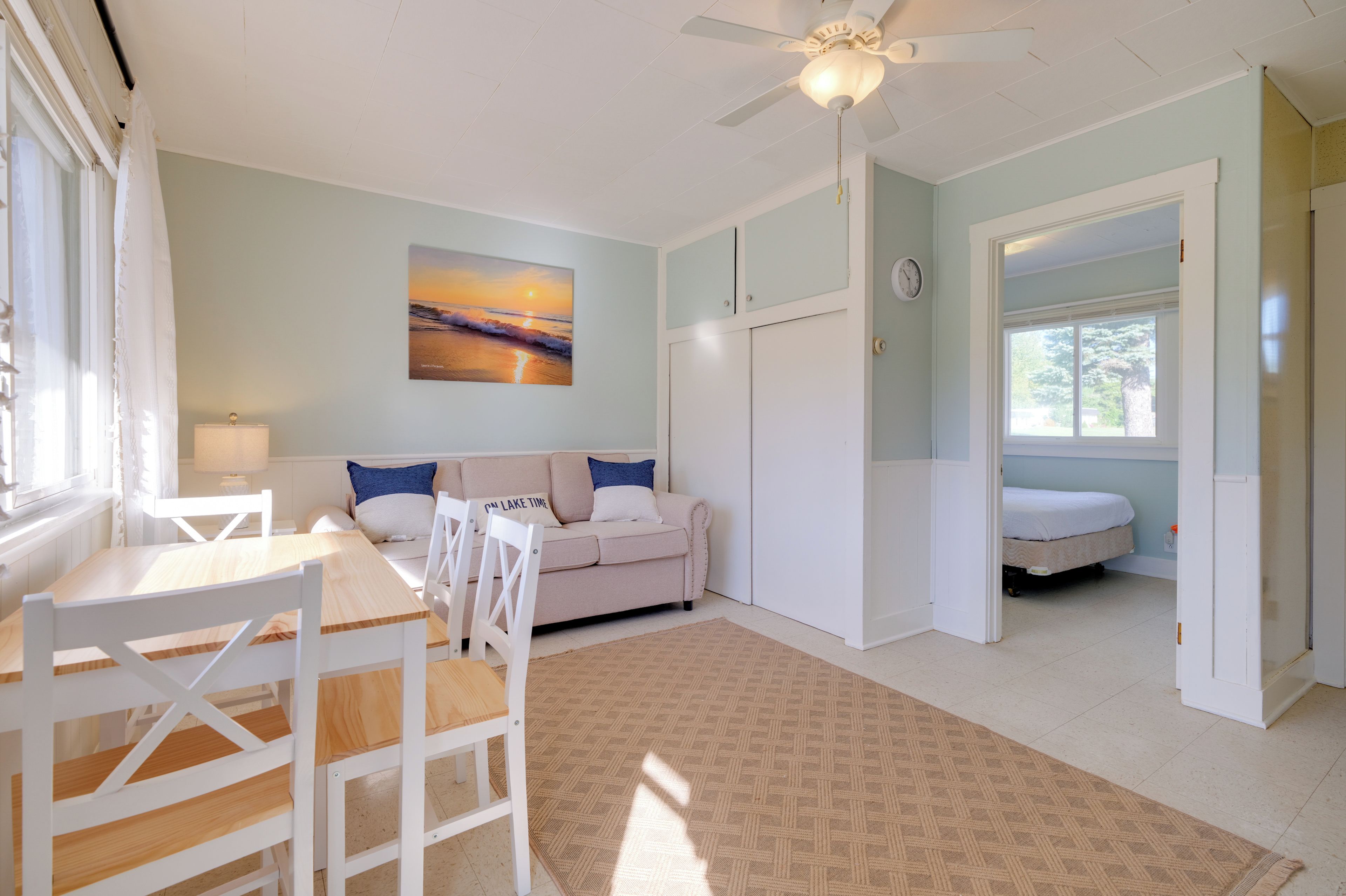 A room with a dining table, chairs, a sofa, a bed, a ceiling fan, a painting, and a window.