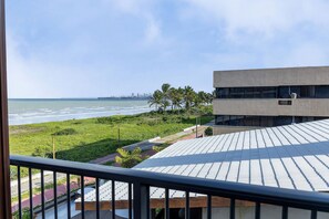 Blick von einem Balkon auf Strand, eine Rasenfläche und ein modernes Gebäude mit flachem Dach.