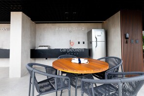 Un área de comedor exterior moderna con sillas de mimbre y una mesa de madera, un refrigerador y un letrero que dice 'Espace Grill 2' al fondo.