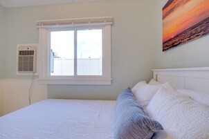 Un dormitorio con cama, ventana, aire acondicionado instalado en la pared y un cuadro de atardecer.