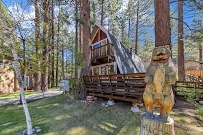 A cabin with a sloped roof, surrounded by tall trees, and a wooden bear sculpture in the foreground. - 1627 Andy Jo Circle 2 Bedroom Cabin (South Lake Tahoe)