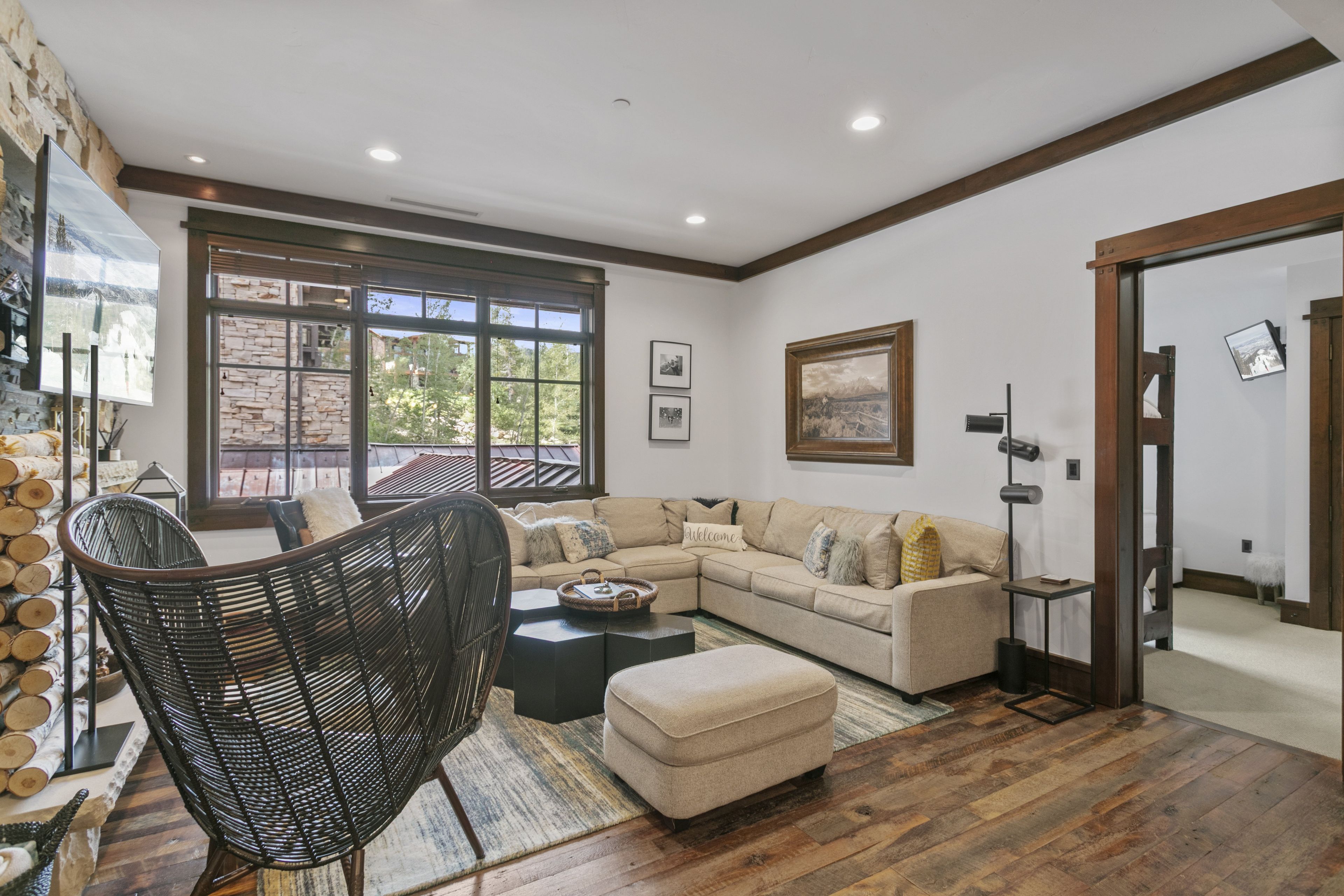A modern living room with a sectional sofa, a wicker chair, and a coffee table. There is a large window with a view of trees and a stone wall.