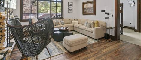 A modern living room with a sectional sofa, a wicker chair, and a coffee table. There is a large window with a view of trees and a stone wall.