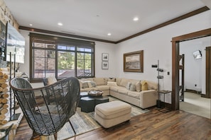 A modern living room with a sectional sofa, a wicker chair, and a coffee table. There is a large window with a view of trees and a stone wall. - 201 - Two Bedroom Arrowleaf 2 Condo (Park City)
