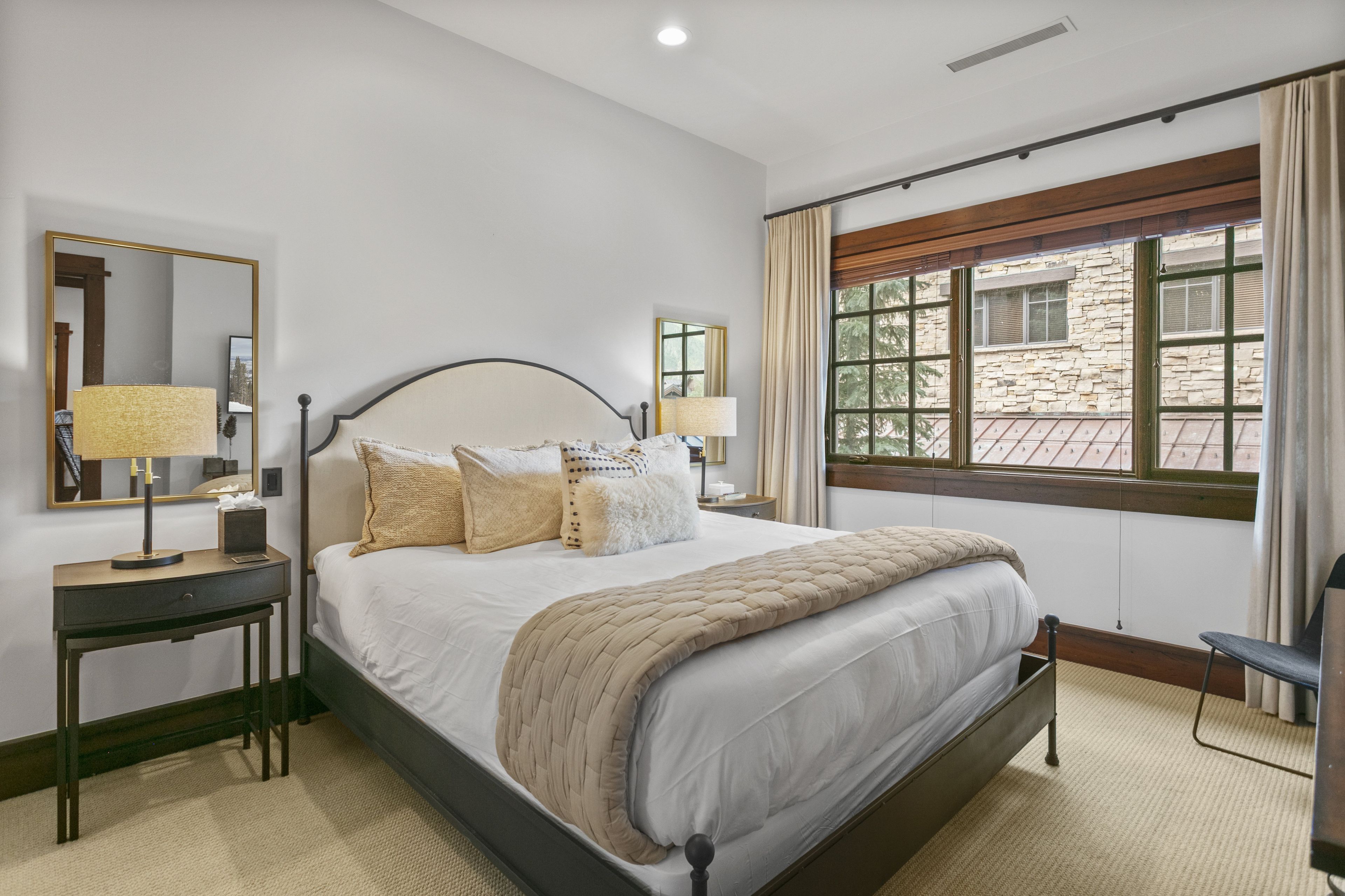 A neatly arranged bedroom with a large bed, bedside tables, a mirror, and a window with a view of a stone building.