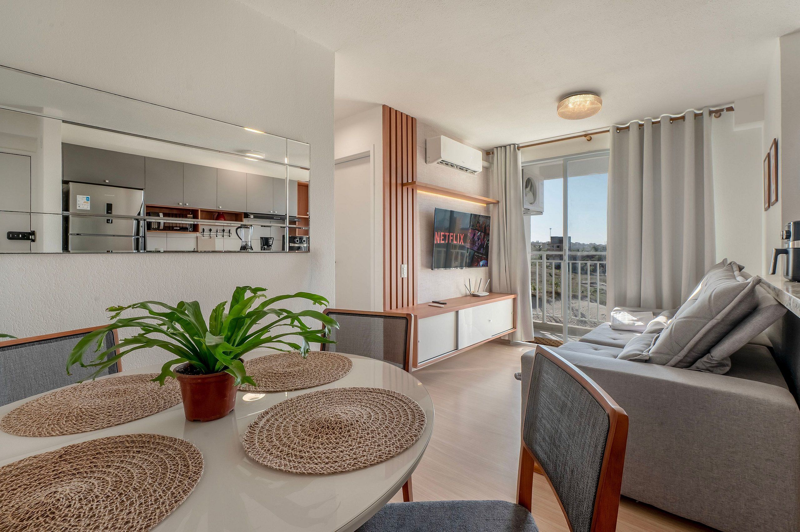 A modern living room with a grey sofa, a dining area with a round table and woven placemats, a kitchen with stainless steel appliances, and a balcony with a view.