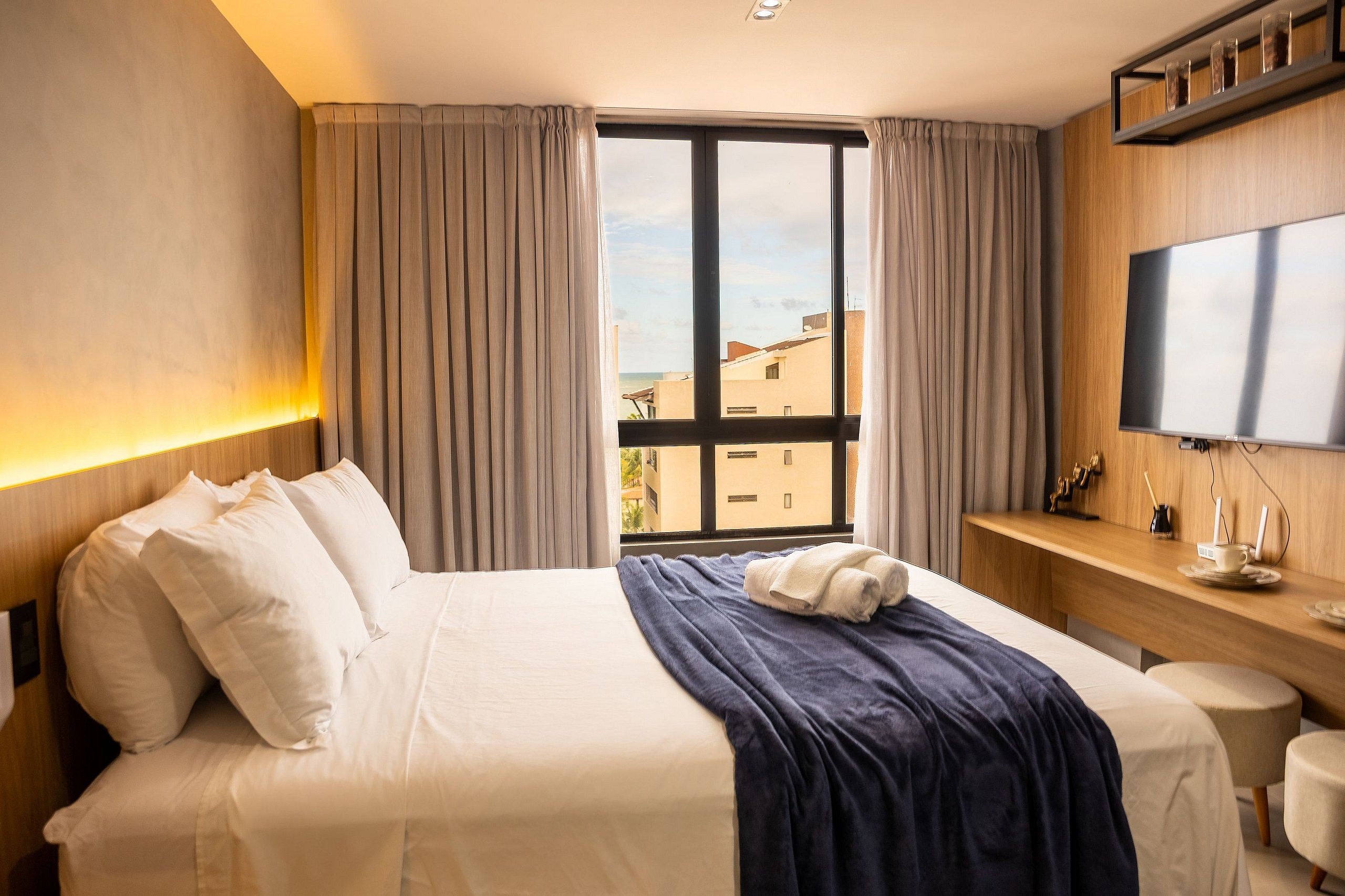 A hotel room with a bed, a TV, a desk with a computer, and a window with curtains.