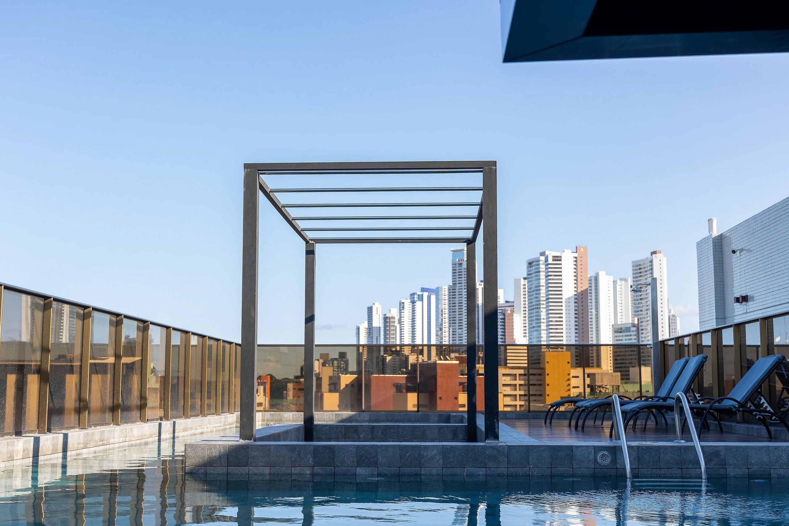 Una piscina sul tetto con ringhiera in vetro, poltrone relax e skyline urbano sullo sfondo.