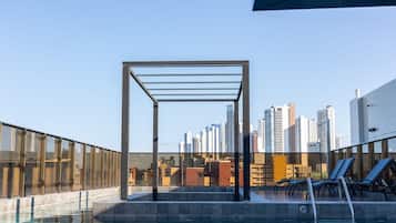 Una piscina sul tetto con ringhiera in vetro, poltrone relax e skyline urbano sullo sfondo.