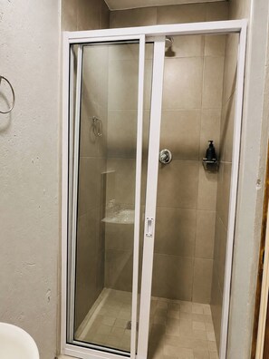 A glass-enclosed shower stall with a white door, beige tiles, a handheld showerhead, and a soap dispenser. - Charming Studio Apartment in Central Randburg (Johannesburg)