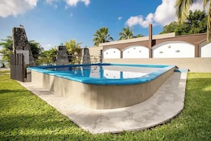 A swimming pool with a curved edge, surrounded by a grassy area and a decorative wall with a stone sculpture. - Palms Reef - Peaceful Condo with Pool & Steps to Beach (Cozumel)