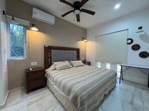 Une chambre à coucher comprenant un lit, des tables de chevet, un ventilateur de plafond, un climatiseur, une fenêtre et un miroir fixé au mur.