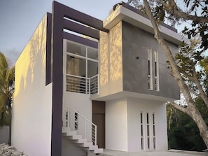 A modern two-story house with a balcony, a staircase, and a tree in the background. - Palms Reef - Peaceful Condo with Pool & Steps to Beach (Cozumel)