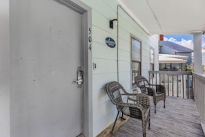 Eine Veranda mit Korbstühlen und einem Willkommensschild, eine Tür mit Hausnummer und Blick auf ein Nachbargebäude.