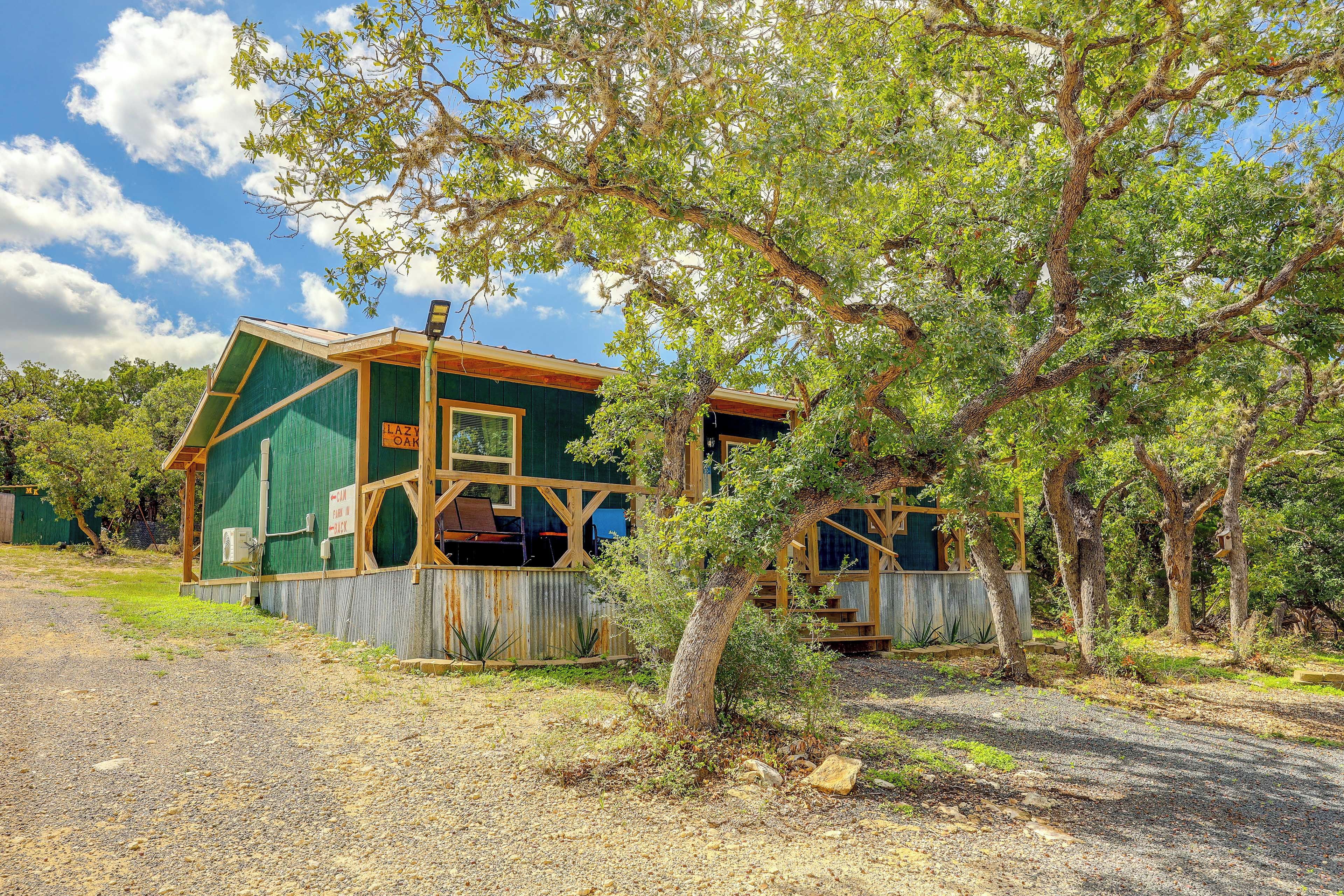 A rustic cabin with a green exterior, a wooden porch, and a gravel driveway.