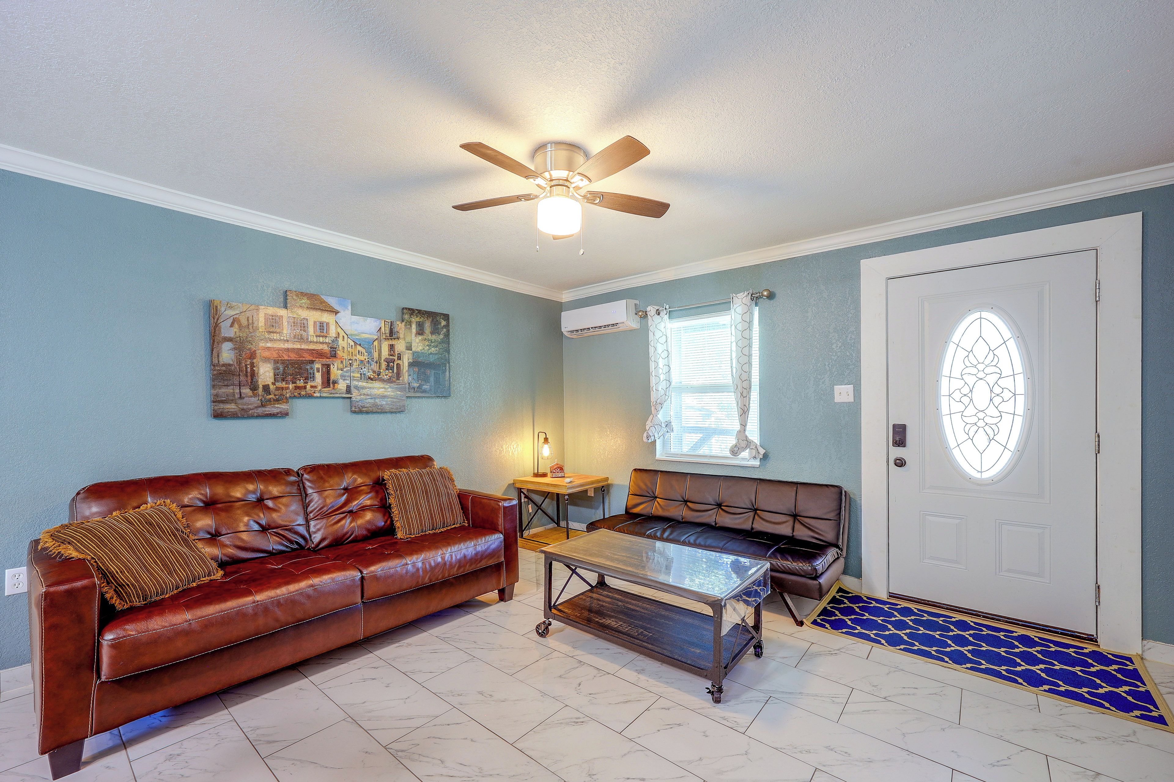 A living room with a brown leather sofa, a glass coffee table, a ceiling fan, and a door with a stained glass window.