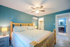 Quarto com cama, luminárias laterais, ventilador de teto e banheiro visível por uma porta aberta.