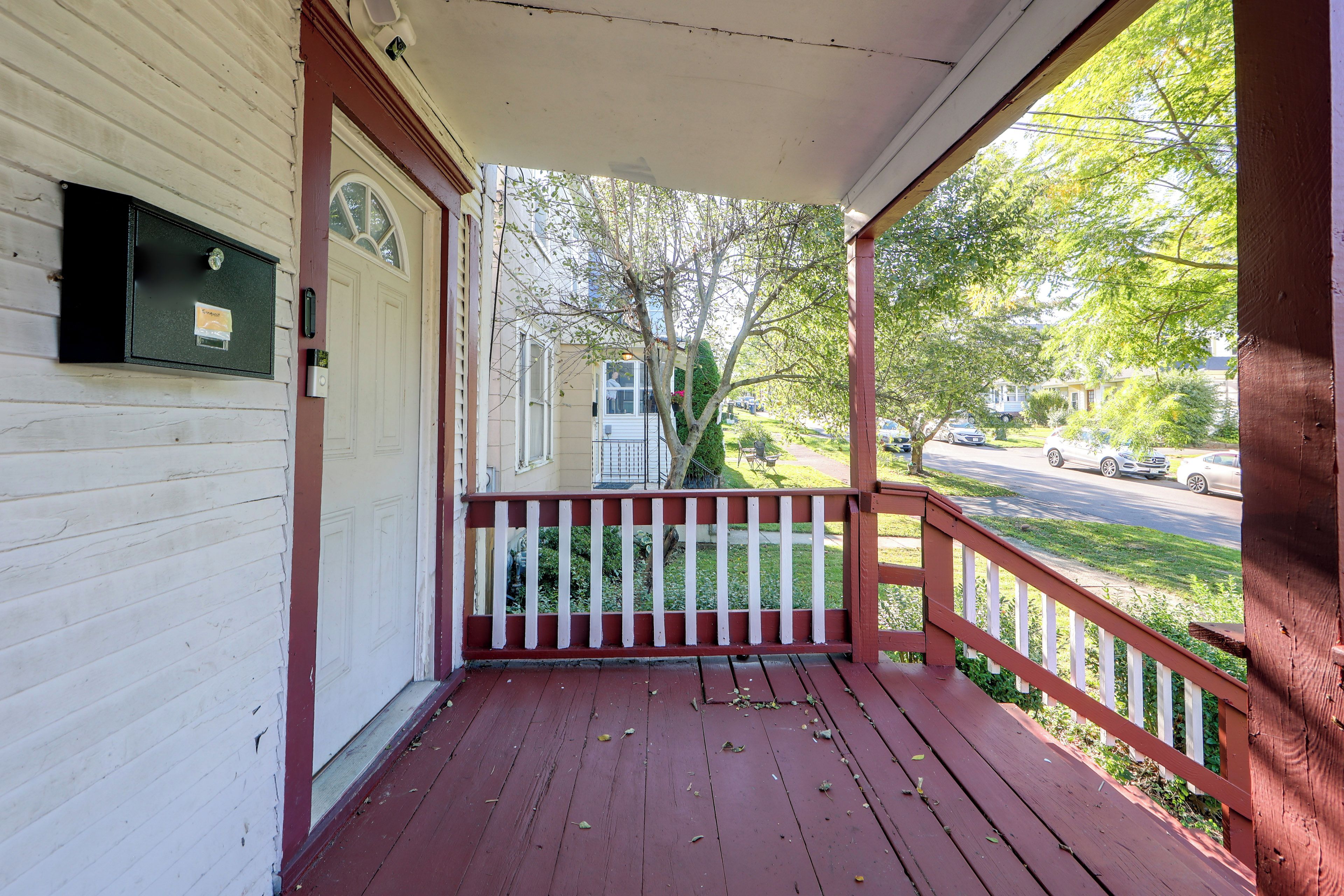 A wooden porch with a red railing, a mailbox, and a view of a street with cars and trees.