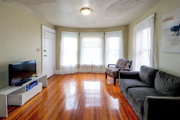 A living room with a flat-screen TV, a sofa, a patterned armchair, and a window with sheer curtains. - Walk to Rosamond Gifford Zoo & Dining! Cozy Unit (Syracuse)