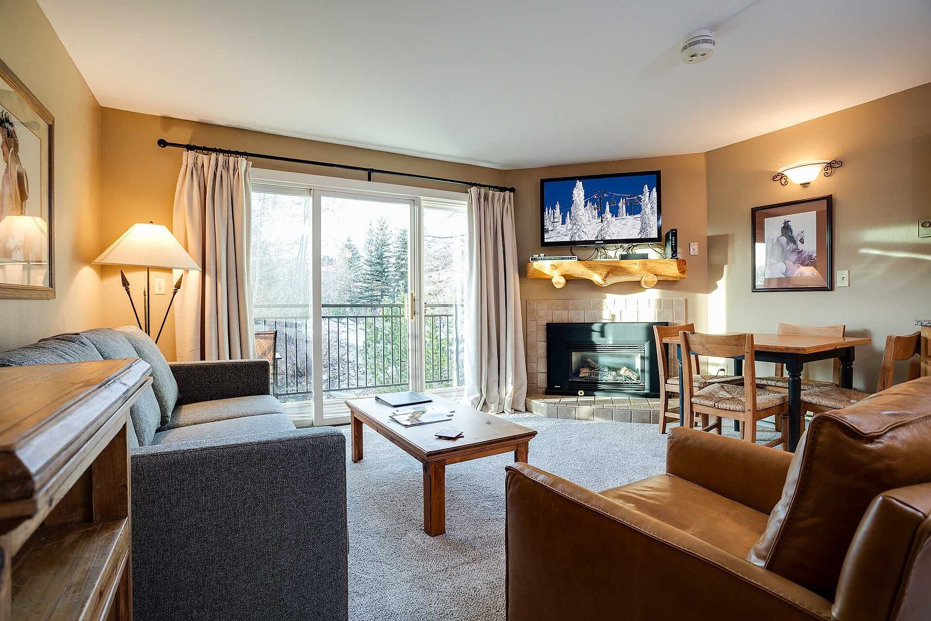 A living room with a sofa, armchair, coffee table, fireplace, television, and a balcony with curtains.