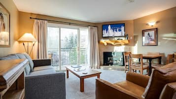 A living room with a sofa, armchair, coffee table, fireplace, television, and a balcony with curtains.
