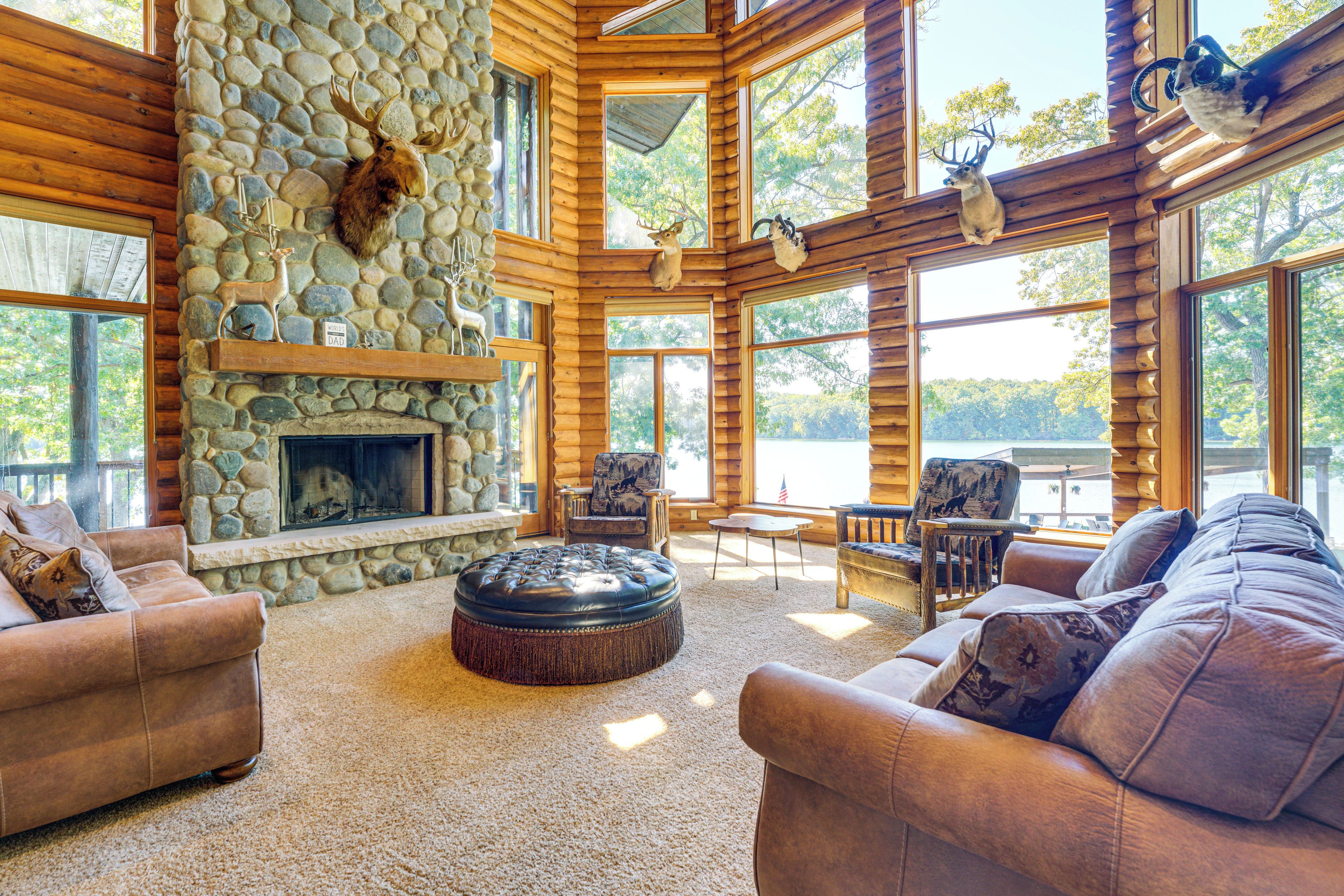 A log cabin interior with a stone fireplace, leather furniture, and large windows overlooking a forest.