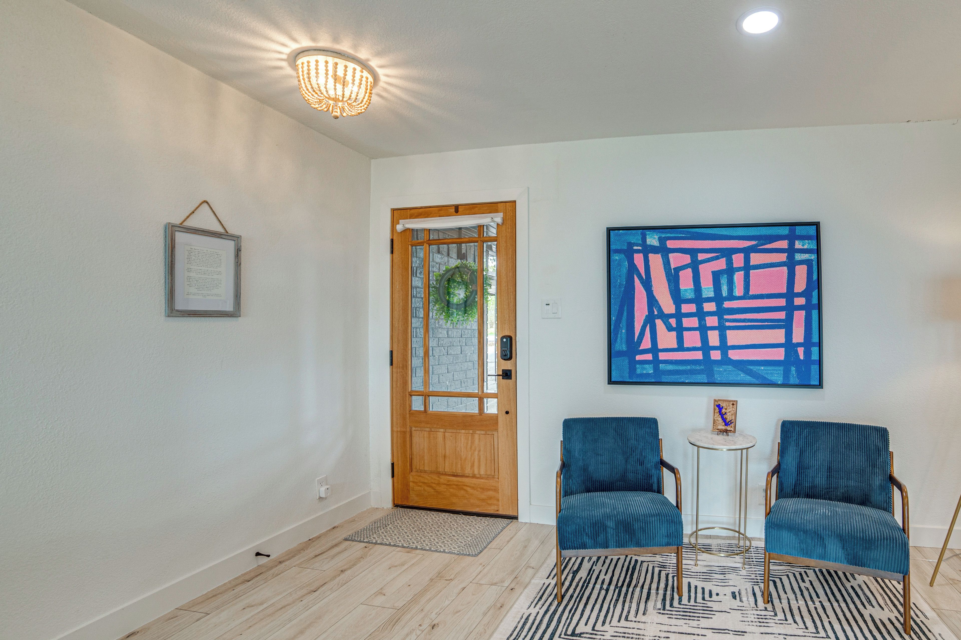 A room with a wooden door, a framed picture, two blue chairs, a small table, and a blue abstract painting on the wall.