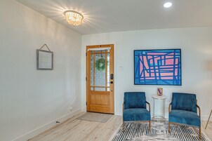 Una habitación con puerta de madera, un cuadro enmarcado, dos sillas azules, una mesita y un cuadro abstracto azul en la pared.