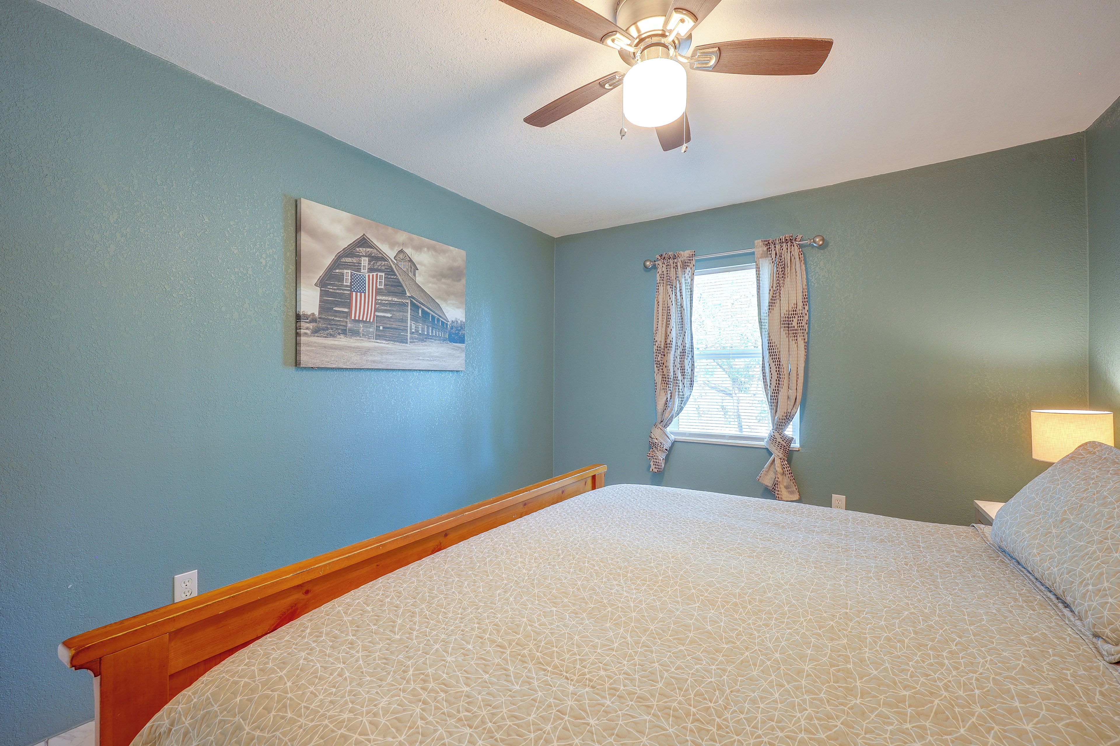 A bedroom with a bed, a ceiling fan, a window with curtains, and a wall painting.