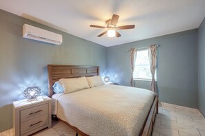 Quarto com cama, mesa de cabeceira, ventilador de teto, aparelho de ar condicionado e janela com cortinas.