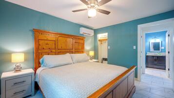 A bedroom with a wooden headboard, a ceiling fan, a bed with patterned bedding, bedside tables with lamps, and a bathroom visible through an open door.