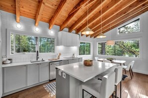 A modern kitchen with white cabinetry, a central island, and large windows offering views of trees. - 3739 Terrace Drive 2 Bedroom Home (South Lake Tahoe)