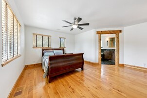 Un dormitorio con piso de madera, una cama, un ventilador de techo y vista a la cocina.