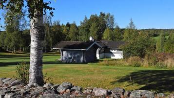 A log cabin with a sloped roof, surrounded by a stone wall, set in a grassy area with trees in the background.