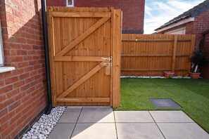 A wooden gate with a metal latch, a brick wall, a grassy yard, and a potted plant. - House Sleeps 7 With Garden (Wolverhampton)