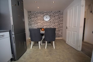 A compact kitchen with a refrigerator, a clock on the wall, and two chairs around a table. - House Sleeps 7 With Garden (Wolverhampton)