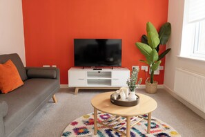 A living room with a sofa, a television on a white stand, a coffee table with decorative items, a potted plant, and a window with blinds. - House Sleeps 7 With Garden (Wolverhampton)