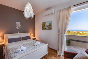 A bedroom with a bed, bedside lamps, a dreamcatcher, a painting, and a view of the landscape. - Villa Kirianna by Villa Plus (Kirianna)