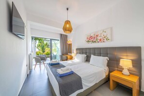 Habitación de hotel con cama, mesitas de noche, televisor, ventana con vistas a vegetación y lámpara colgante.