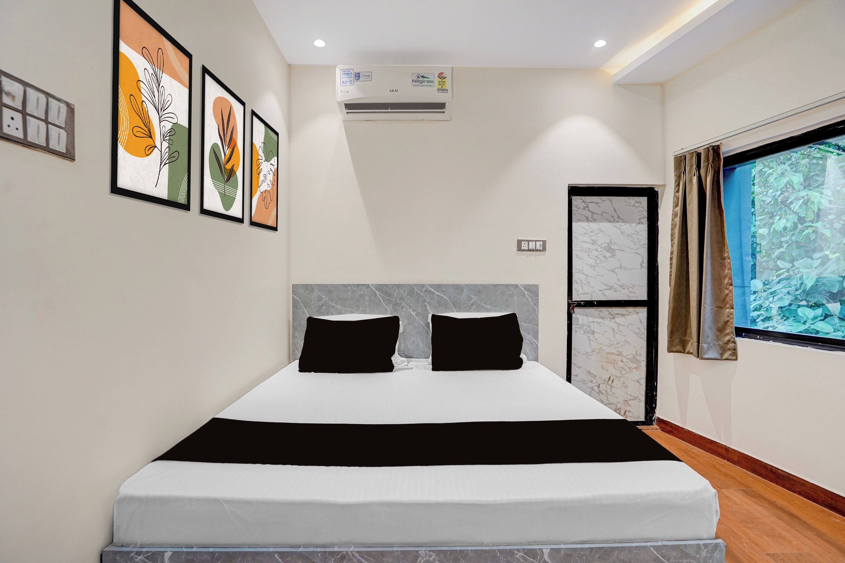 Una habitación de hotel moderna con una cama grande, obras de arte enmarcadas en la pared, un equipo de aire acondicionado y una ventana con vistas a zonas verdes.