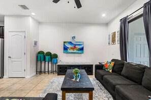 Ein modernes Wohnzimmer mit einem schwarzen Sofa, einem Couchtisch und einem an der Wand befestigten Fernseher. Drei Blumentöpfe stehen auf Beistelltischen, und es gibt eine dekorative Wandkunst mit Blättern.