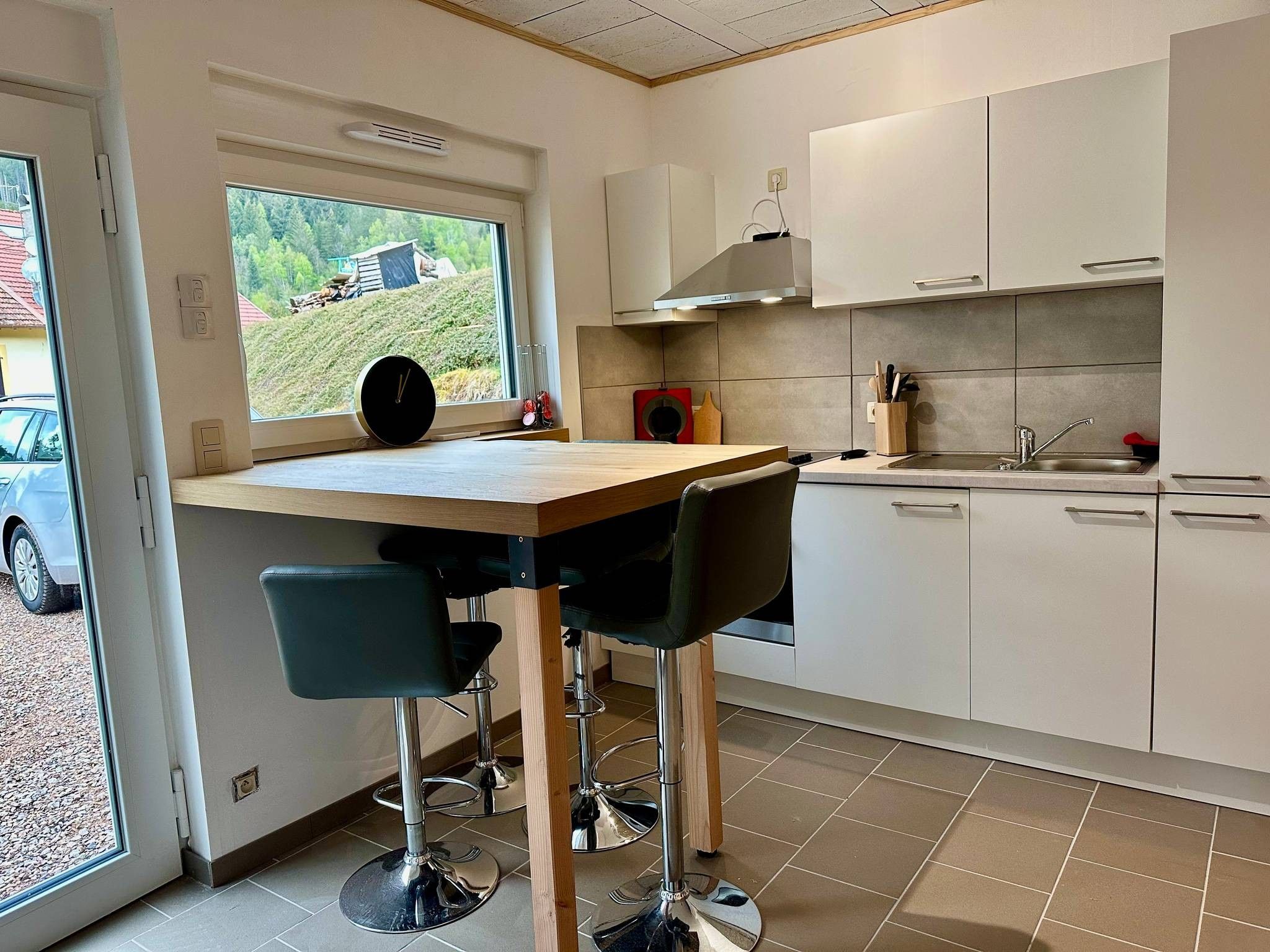 Une cuisine moderne avec un îlot central en bois, deux tabourets de bar, une fenêtre donnant sur une maison et des arbres, et des meubles de rangement blancs.